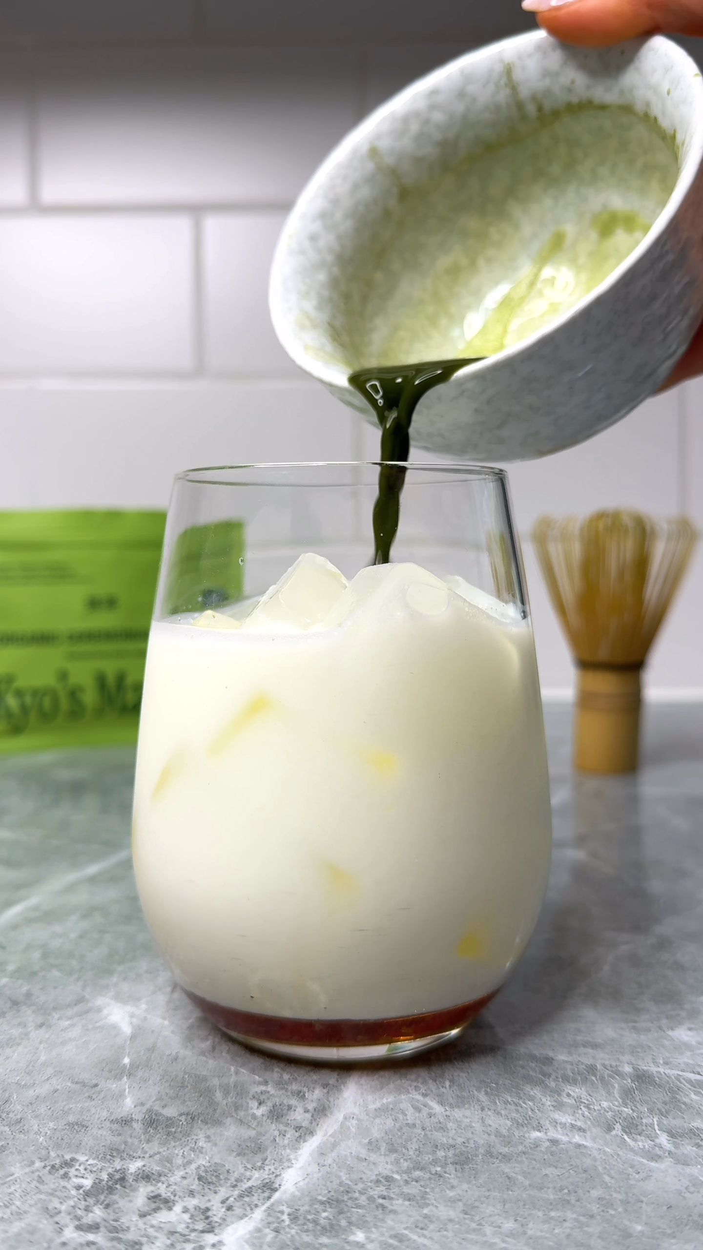 Pouring green liquid from a small bowl into a glass of white liquid on a gray surface.